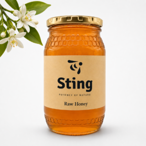 Amber glass honey jar with a gold lid and beige label reading 'Sting Raw Honey' beside a small white flower and green leaves